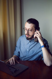 Businessman with a beard and glasses is sitting at a laptop at a wooden table. talking on the phone