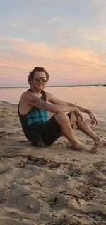 Portrait of man sitting on shore at beach against sky during sunset