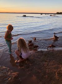 Sisters playing with birds on shore at beach against sky during sunset