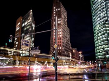 Light trails on city street by buildings against sky at night