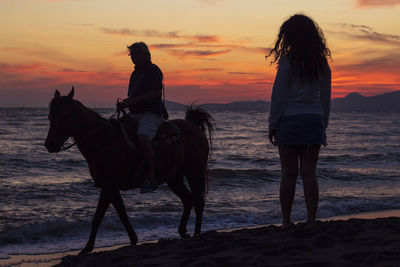 Silhouette woman standing by friend riding horse at beach against sky during sunset