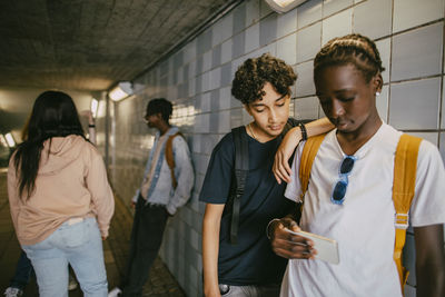 Teenage boy standing with hand on shoulder of male friend using smart phone in tunnel