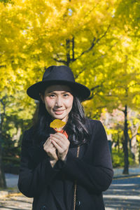 Portrait of woman holding leaf while standing against trees in park during autumn