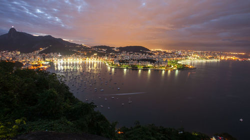 Illuminated cityscape by sea against sky during sunset