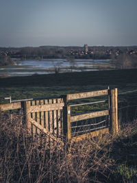 Wooden fence on field by lake against sky