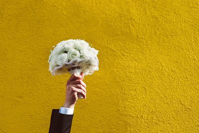 Cropped hand of bridegroom holding bouquet against yellow wall