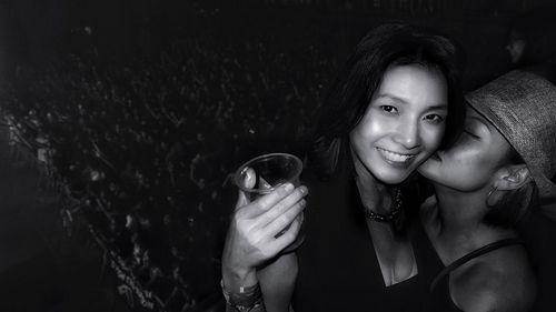 Portrait of smiling young woman holding drinking glass while friend kissing her at night