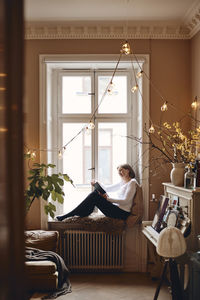 Side view of elderly woman reading book while sitting on window sill
