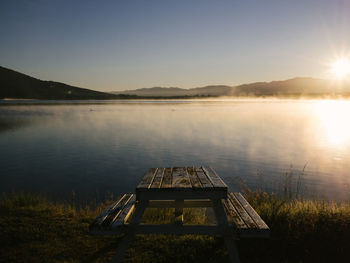 Picnic table at lakeshore against sky during sunrise