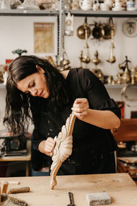 Skilled female artisan polishing decorative carved wooden molding with abrasive paper while working in professional workshop