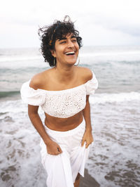 Positive ethnic female in white stylish outfit and with curly hair standing on seashore and laughing with closed eyes