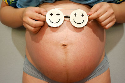 Midsection of pregnant woman in nightwear with smiley face eyeglasses standing against wall