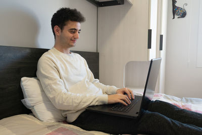 Young caucasian man working on his laptop in the bed, with a casual pajamas and smiling