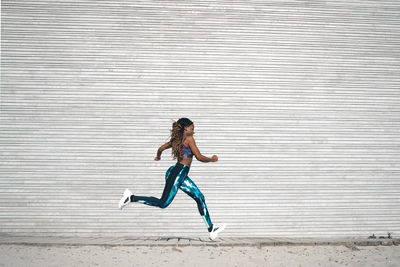 Full body side view of slim african american female with flying long dreadlocks wearing tight sportswear jogging on walkway near metal wall during cardio workout