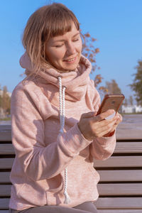 Smiling young woman using mobile phone wile standing against sky