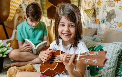 Portrait of smiling little girl playing ukulele while boy reading book on handmade teepee at home