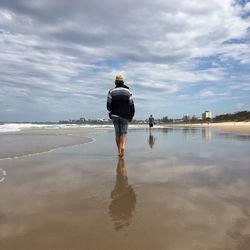 Rear view of men walking at beach against cloudy sky