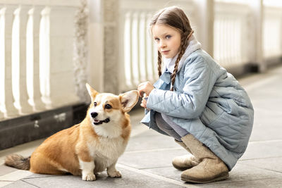 Portrait of a toddler girl in a warm blue coat with a corgi dog in the park. 