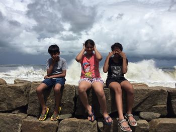 Full length of friends posing while sitting on retaining wall against sea