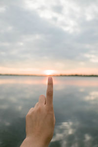 Optical illusion of finger touching sun by lake against sky during sunset