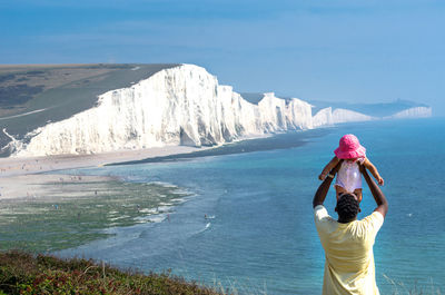 Rear view of father playing with daughter by sea against mountains and sky