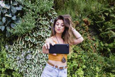 Beautiful woman taking selfie from mobile phone while standing against plants at park