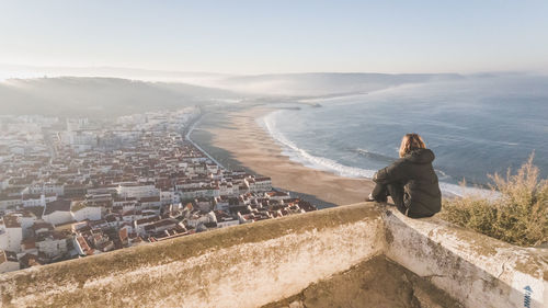 Rear view of woman relaxing on retaining wall against city by sea