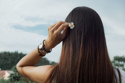 Rear view of woman putting flower in hair against sky