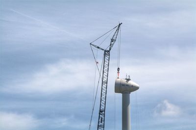 Low angle view of crane and windmill against cloudy sky