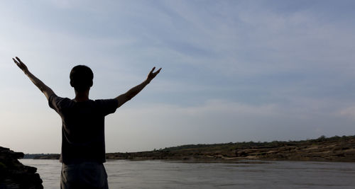 Rear view of man with arms outstretched standing by lake against sky