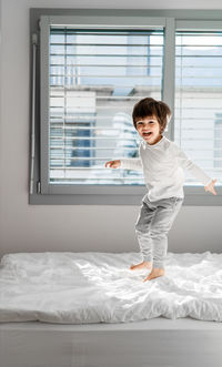 Full length of cute boy standing on bed by window at home