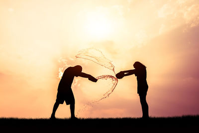 Silhouette friends splashing water with containers while standing on field against orange sky