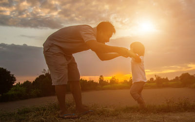 Side view of father and son standing on shore against sky during sunset