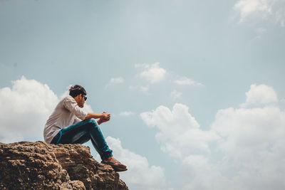 Side view of young man sitting on rock against sky