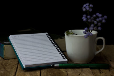 Close-up of flowers in cup by spiral notebook on wooden table against black background