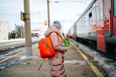 Girl on the platform of the station with a red backpack and a bouquet of tulip flowers