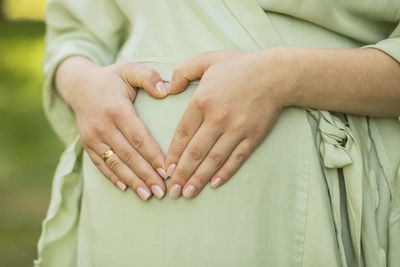 Cropped pregnant woman holds hands in heart shape on big belly, green trees, meadow on background