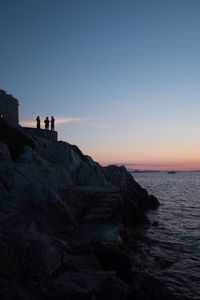 People on rocks by sea against sky during sunset