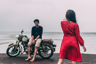 Rear view of woman wearing red dress standing at beach while man sitting on motorcycle in background