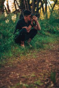 Young man photographing while crouching on field in forest