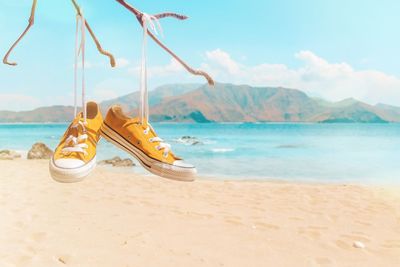 Yellow canvas shoes hanging from branch at beach against sky