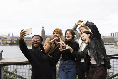 Cheerful female friends making heart gesture while taking selfie through smart phone on vacation