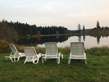 Chairs on grass by lake against sky