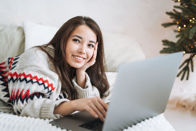 Young asian woman with dark long hair in cozy sweater using laptop on bed christmas at home