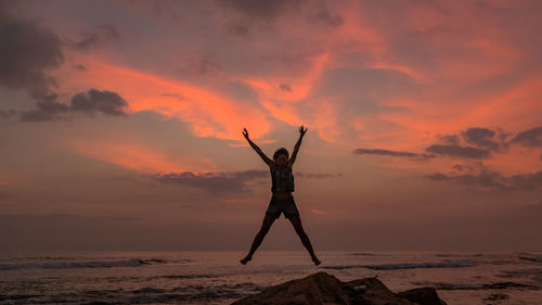 Woman jumping at rocky shore against orange sky