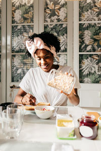 Happy african american lady smiling and adding fresh oatmeal in bowl while sitting at table and having breakfast at home