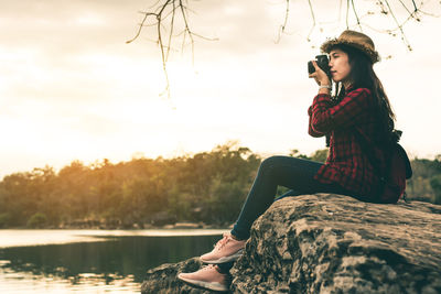 Young woman photographing while sitting over lake against trees