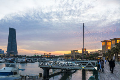 People on harbor by buildings against sky during sunset