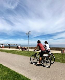 Friends riding bicycle on footpath against cloudy sky