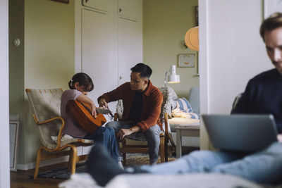 Father teaching daughter to play guitar while sitting on chair at home